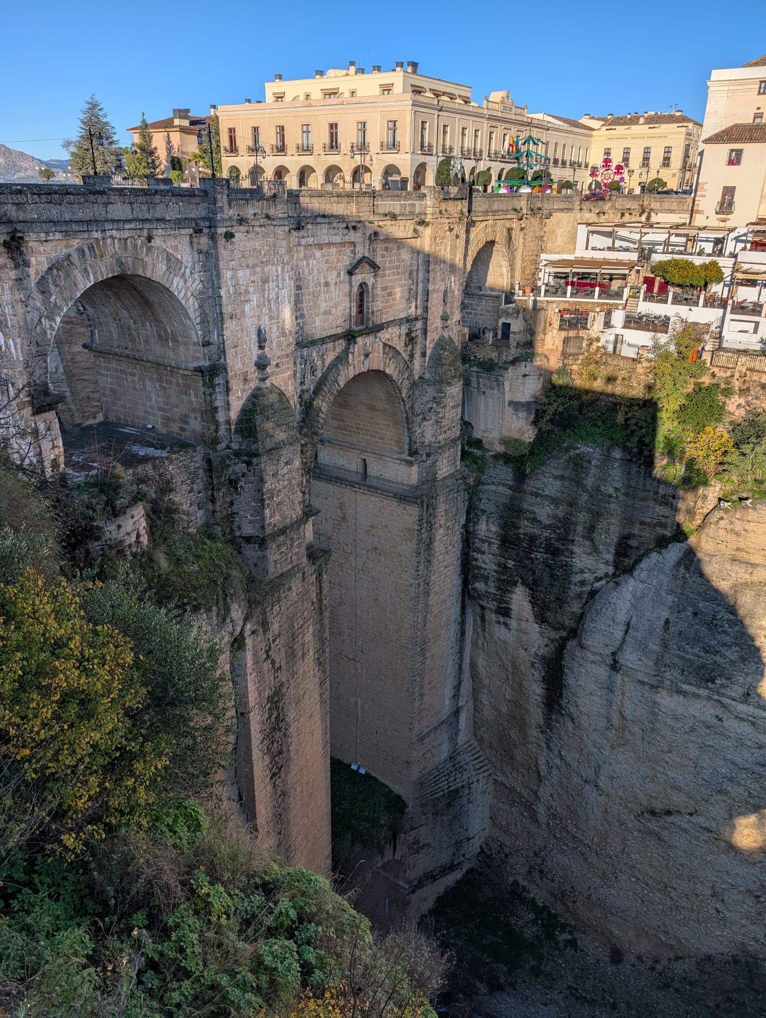 Deze foto toont de Puente Nuevo in Ronda, één van de meest iconische bruggen van Andalusië. De brug overspant de diepe El Tajo-kloof, een spectaculaire ravijn die de oude en nieuwe stad van Ronda van elkaar scheidt. Met een hoogte van ongeveer 98 meter boven de rivier is dit niet alleen een indrukwekkend bouwwerk, maar ook één van de bekendste uitzichtpunten van Zuid-Spanje.

Wat opvalt op de foto is de massieve stenen constructie met de grote centrale boog en daarboven de kleinere openingen. In het midden van de brug bevindt zich zelfs een kamer, die in het verleden onder andere als gevangenis is gebruikt. De gebouwen bovenop de kloof laten goed zien hoe de stad letterlijk op de rand van de afgrond is gebouwd.